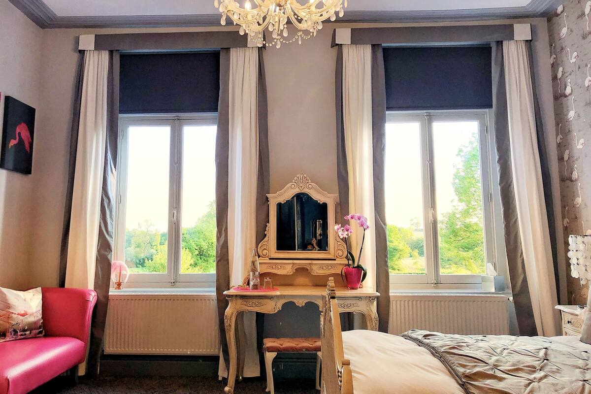 A bedroom with a large window, a chandelier, a vanity with a mirror, a pink chair, and a bed.