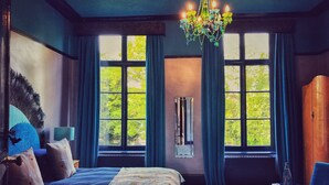 A bedroom with a large bed, two windows with curtains, a chandelier, and a wooden wardrobe. - Maison de Plumes (Heuchin)