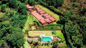 An aerial view of a resort with multiple red-roofed buildings, a swimming pool, and lush greenery surrounding the property. - Hotel Mango Valley (San Roque)