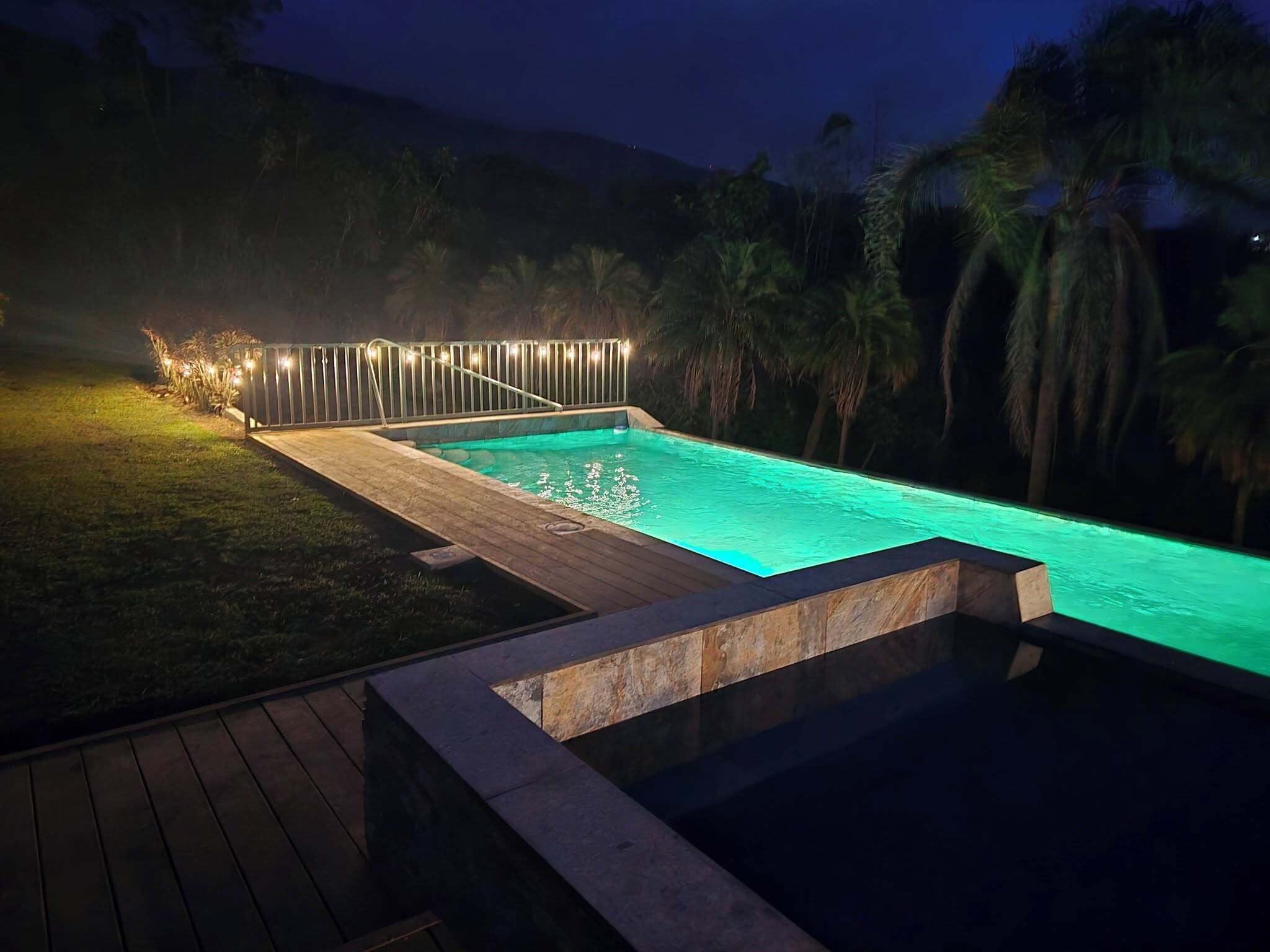 A rectangular swimming pool with a wooden deck and stone edging, surrounded by a fence with string lights.
