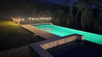 Una piscina rectangular con muelle de madera y borde de piedra, rodeada por una valla con luces de cuerda.