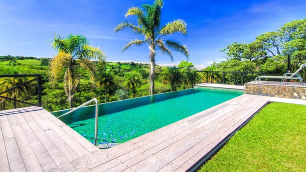 A wooden deck leading to a clear blue pool surrounded by lush greenery and palm trees. - Hotel Mango Valley (San Roque)