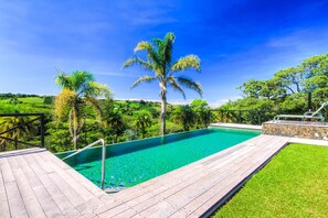 A wooden deck leading to a clear blue pool surrounded by lush greenery and palm trees. - Hotel Mango Valley (San Roque)