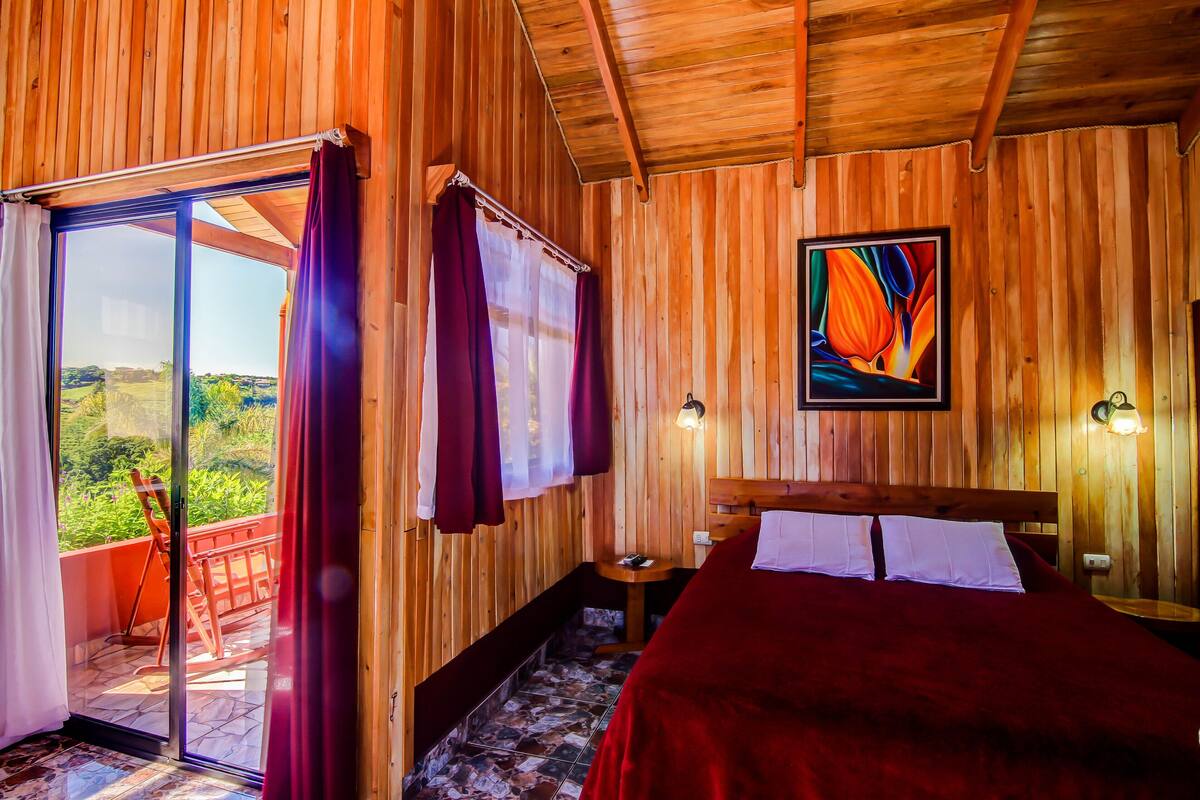 A wooden room with a bed, a painting, and a balcony with a table and chairs.