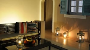 Un salon chaleureux avec un canapé, une table basse sur laquelle se trouvent une bouteille et des fruits, et une fenêtre avec des volets.