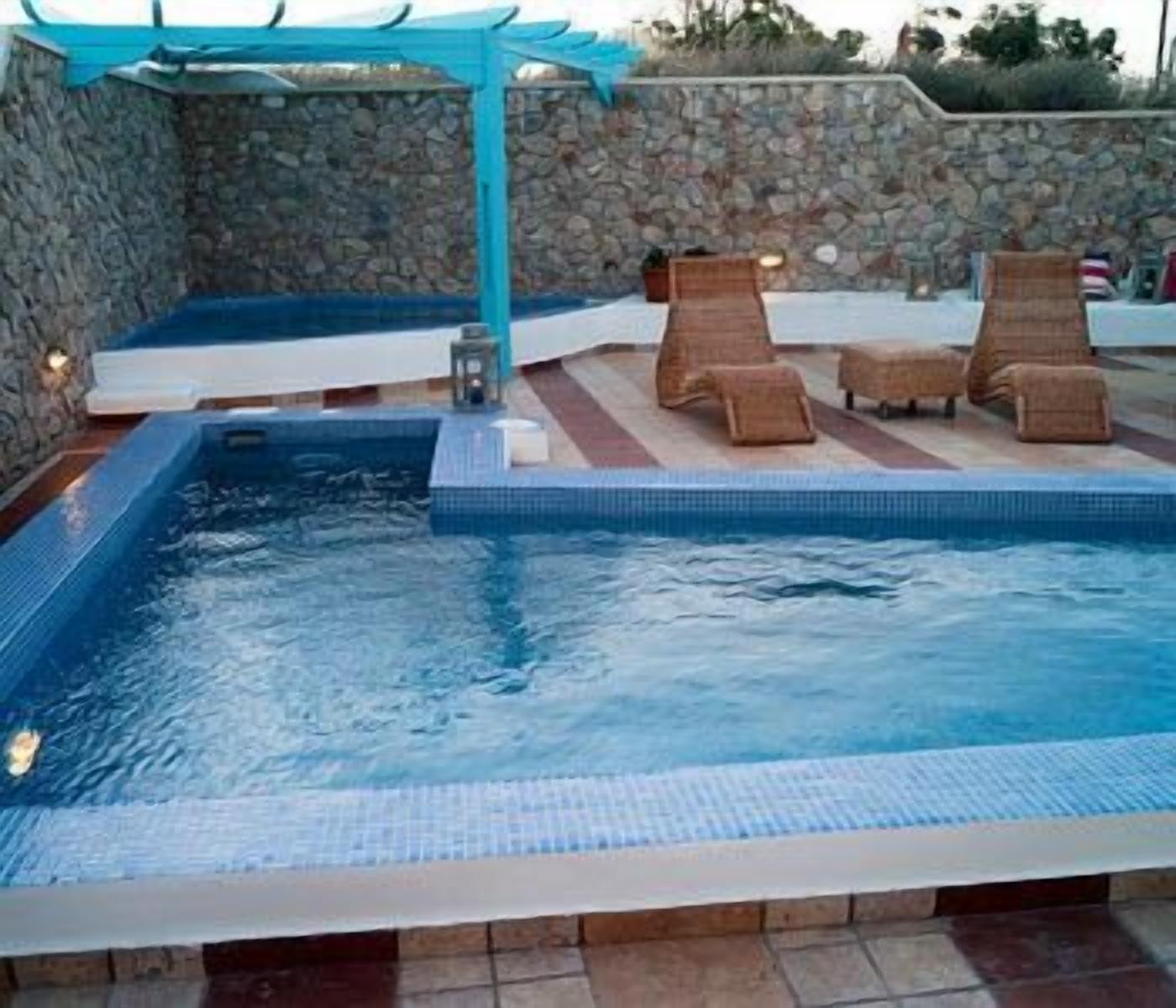 Piscina con pared de piedra y toldo azul. Hay sillones de descanso y una pequeña mesa.