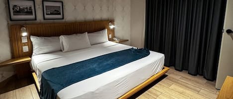 A modern hotel room with a large bed, wooden headboard, two bedside tables, and a dark curtain.