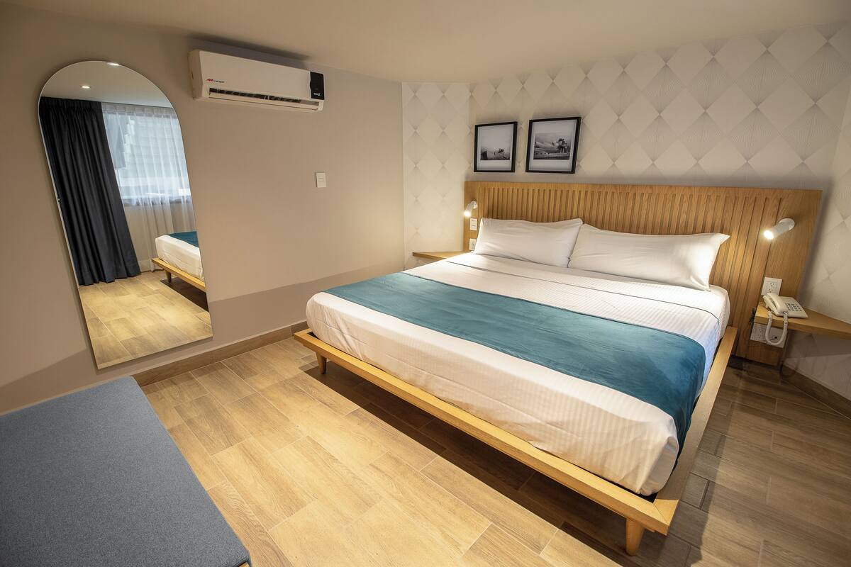 Una habitación de hotel moderna con una cama de madera, una mesita de noche con teléfono, un espejo y un aire acondicionado de pared.