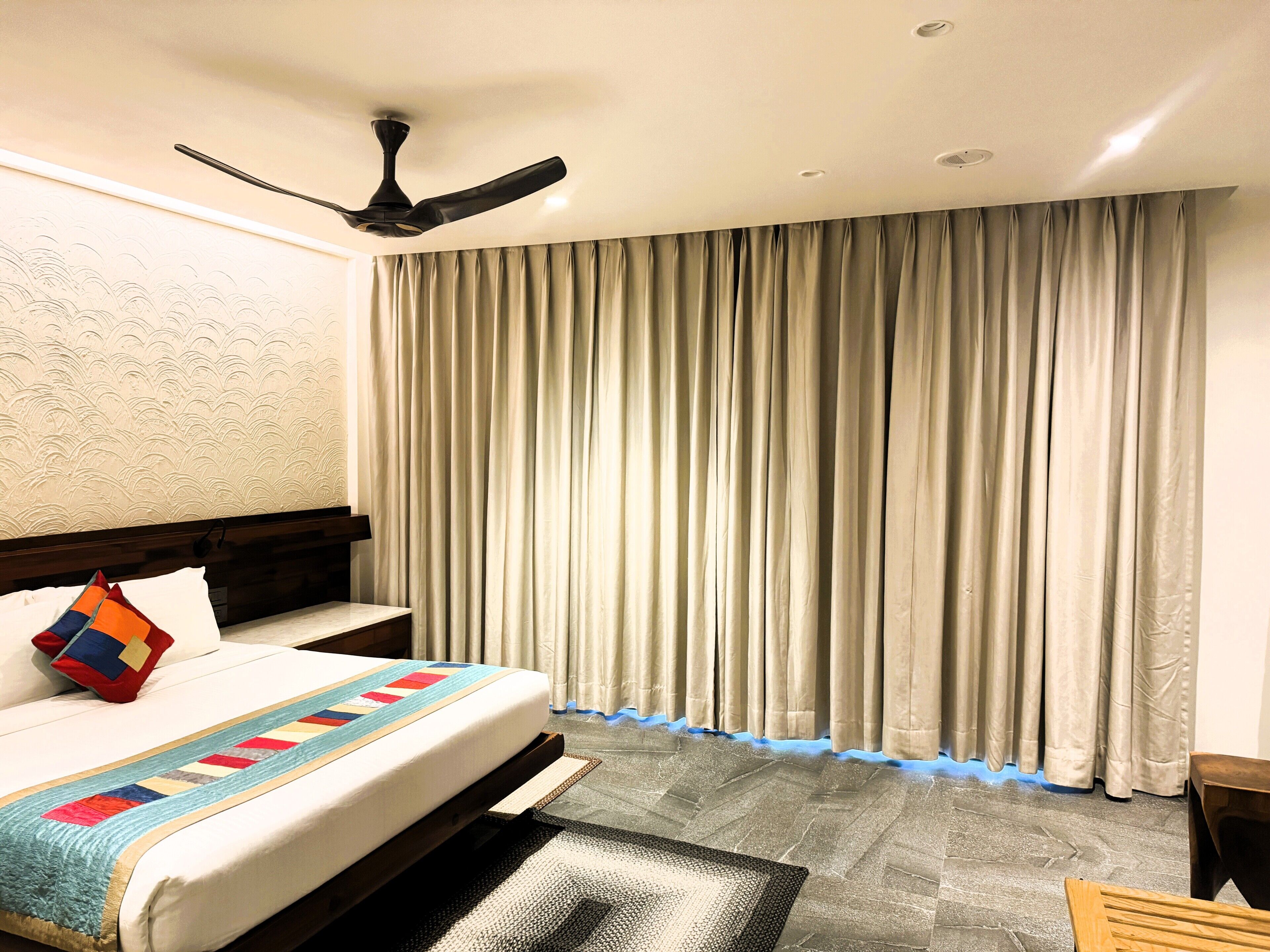 A hotel room with a bed, a ceiling fan, a large window with curtains, and a carpeted floor.