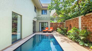 A swimming pool with two orange lounge chairs, a pergola, and a brick wall with plants.