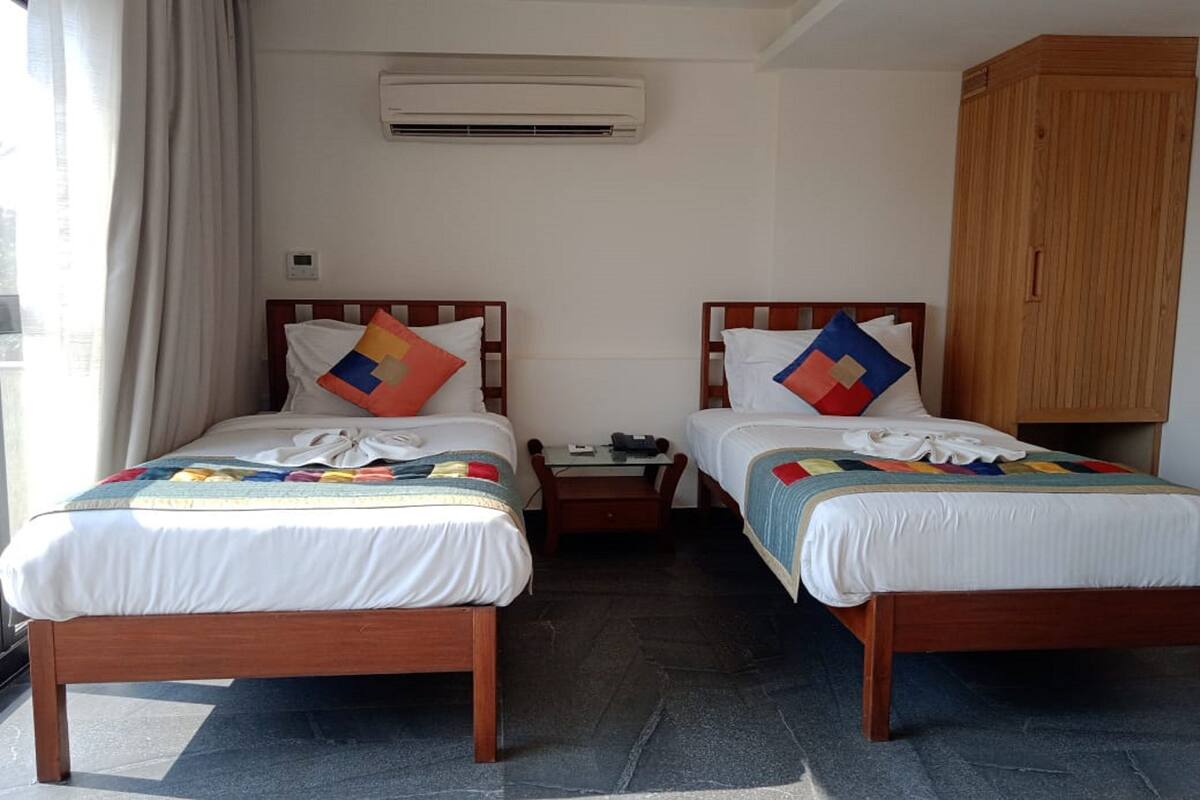 A hotel room with two wooden beds, a small table, an air conditioning unit, and a wardrobe.