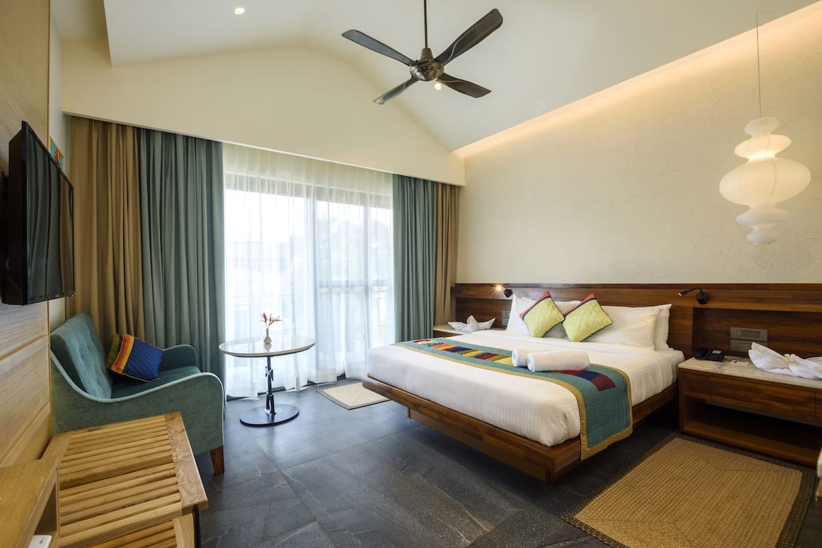 A hotel room with a bed, a sofa, a small table, a ceiling fan, and a TV.