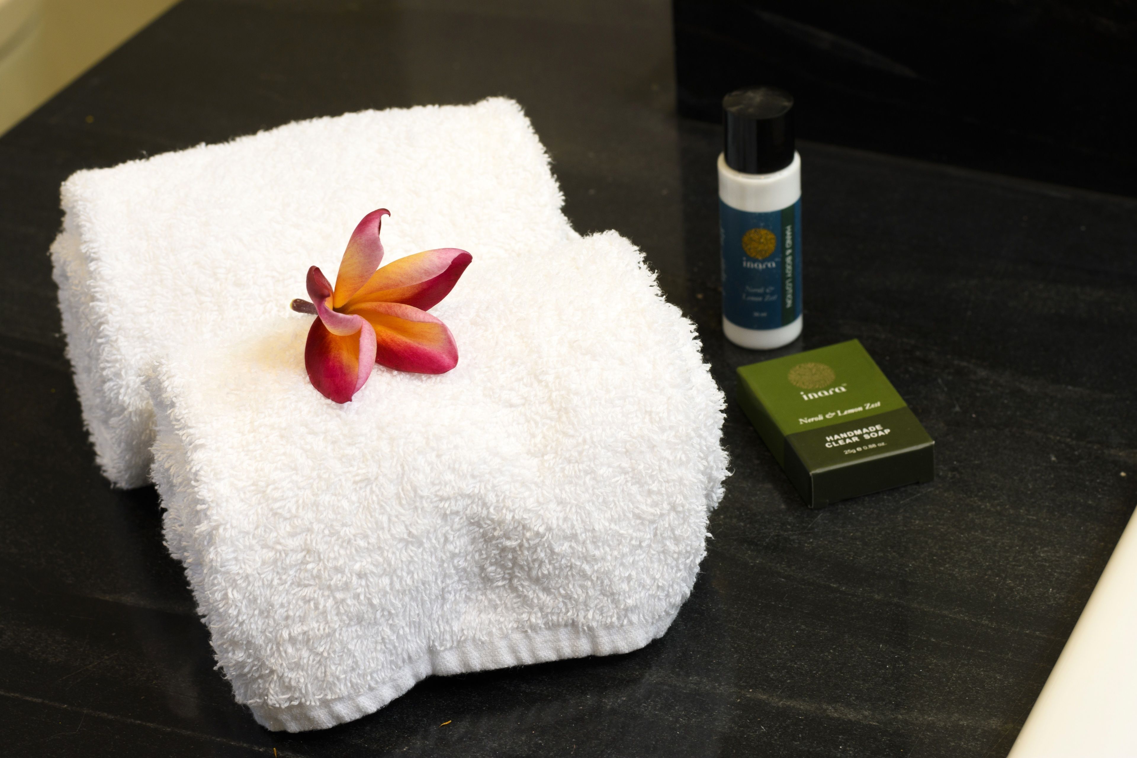 A neatly folded white towel with a flower on it, a bottle of hand soap, and a box of soap on a dark surface.