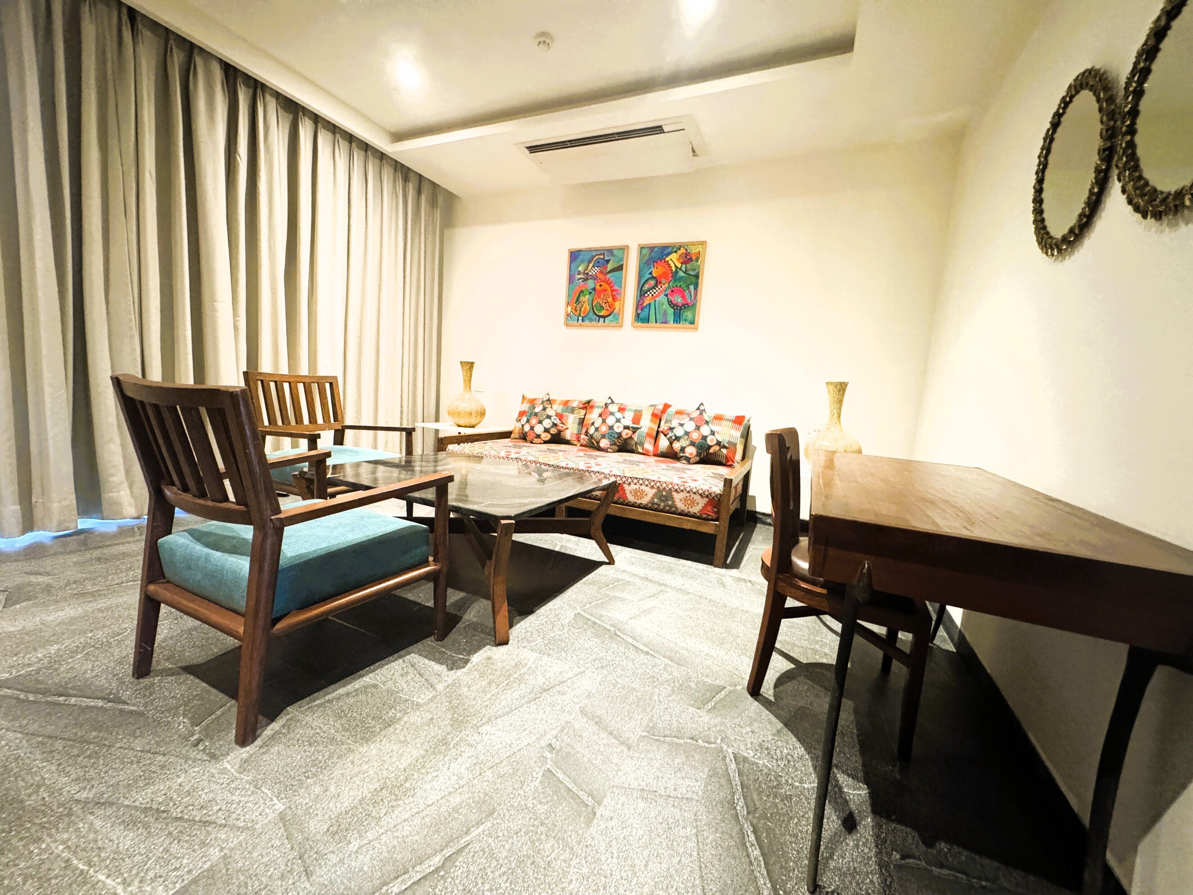 A living room with a sofa, chairs, a coffee table, and a dining table. There are curtains, wall art, and a vase on the table.