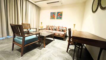 A living room with a sofa, chairs, a coffee table, and a dining table. There are curtains, wall art, and a vase on the table.