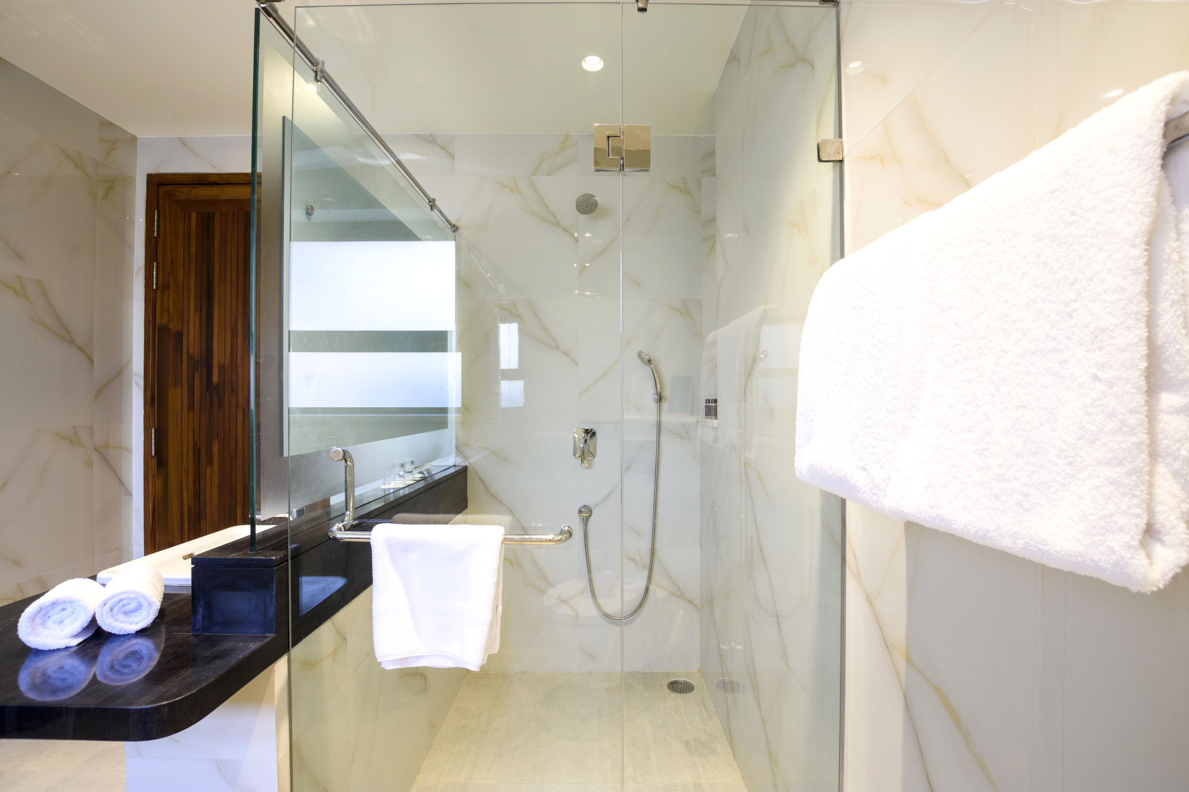 A modern bathroom with a glass-enclosed shower, a marble-tiled wall, a wooden door, and a black countertop with towels.