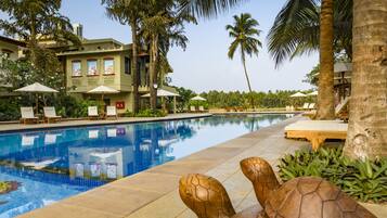 A swimming pool with lounge chairs and umbrellas, surrounded by palm trees and a building with a tiled roof.