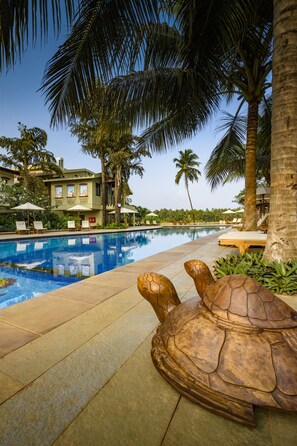 A swimming pool with lounge chairs and umbrellas, surrounded by palm trees and a building with a tiled roof. - Beleza By The Beach (Betalbatim)