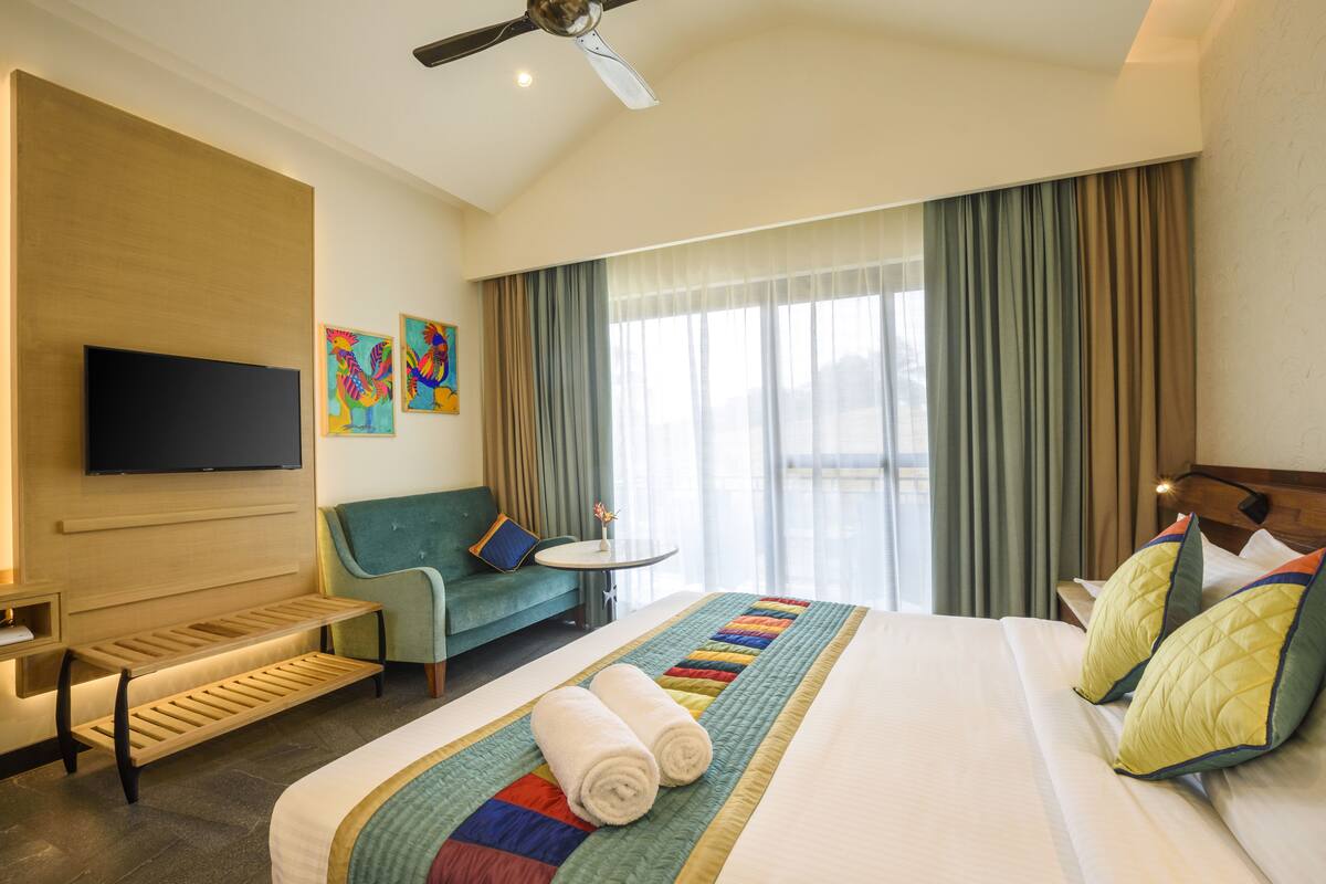A hotel room with a large bed, a sofa, a small table, a TV mounted on the wall, and a window with curtains.