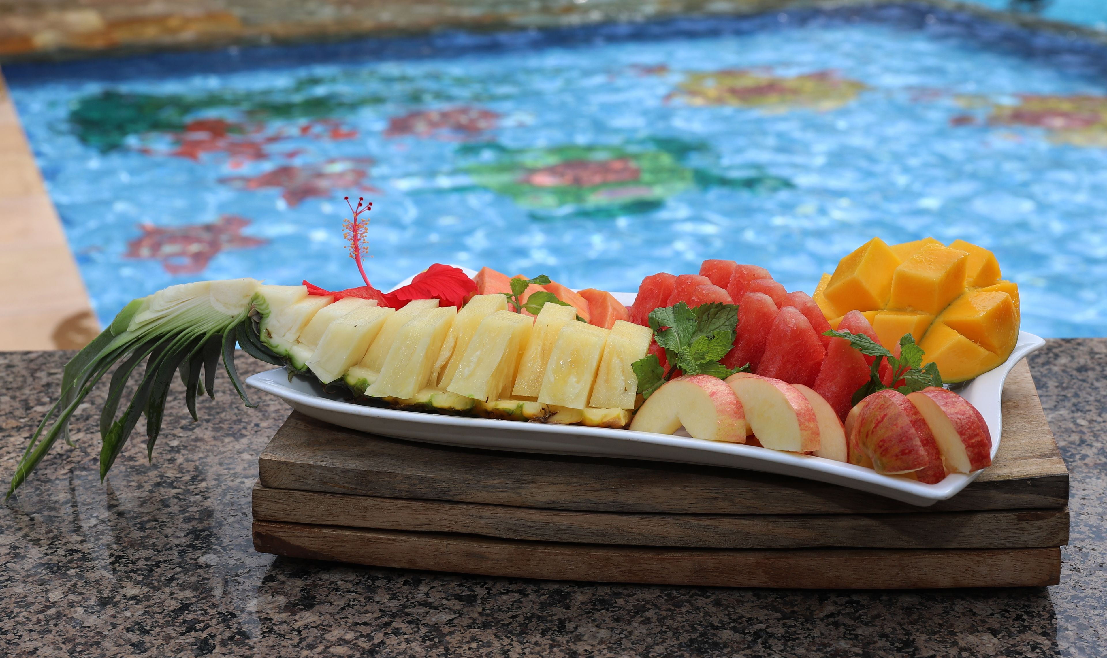 A fruit platter on a wooden board by a pool with a colorful mosaic tile background.