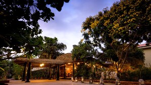 An outdoor area with a thatched-roof pavilion, stone pillars, and a paved walkway. - Beleza By The Beach (Betalbatim)