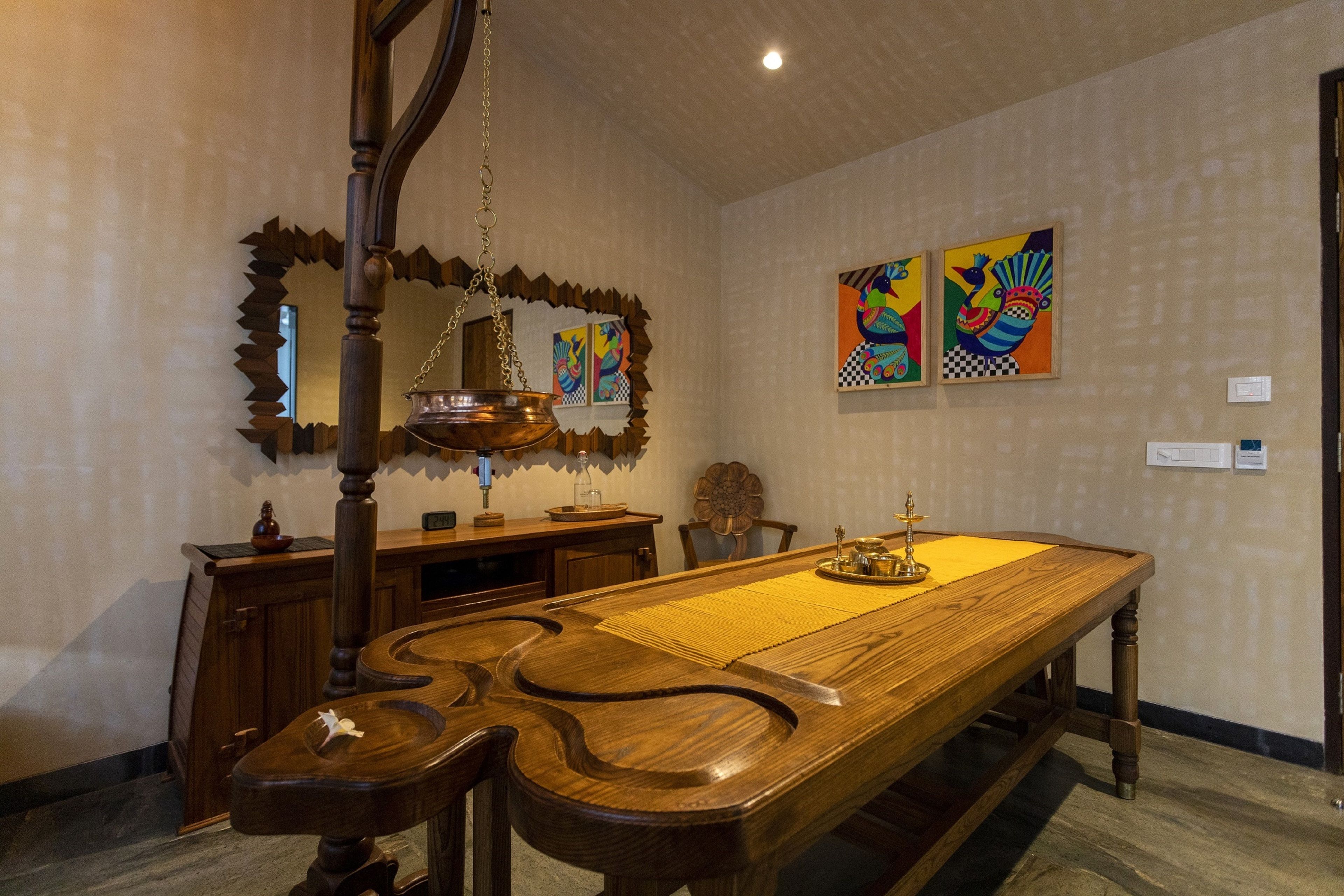 Un comedor con una larga mesa de madera, sillas, una lámpara colgante y dos cuadros coloridos en la pared.