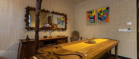 A dining area with a long wooden table, chairs, a hanging light fixture, and two colorful paintings on the wall.