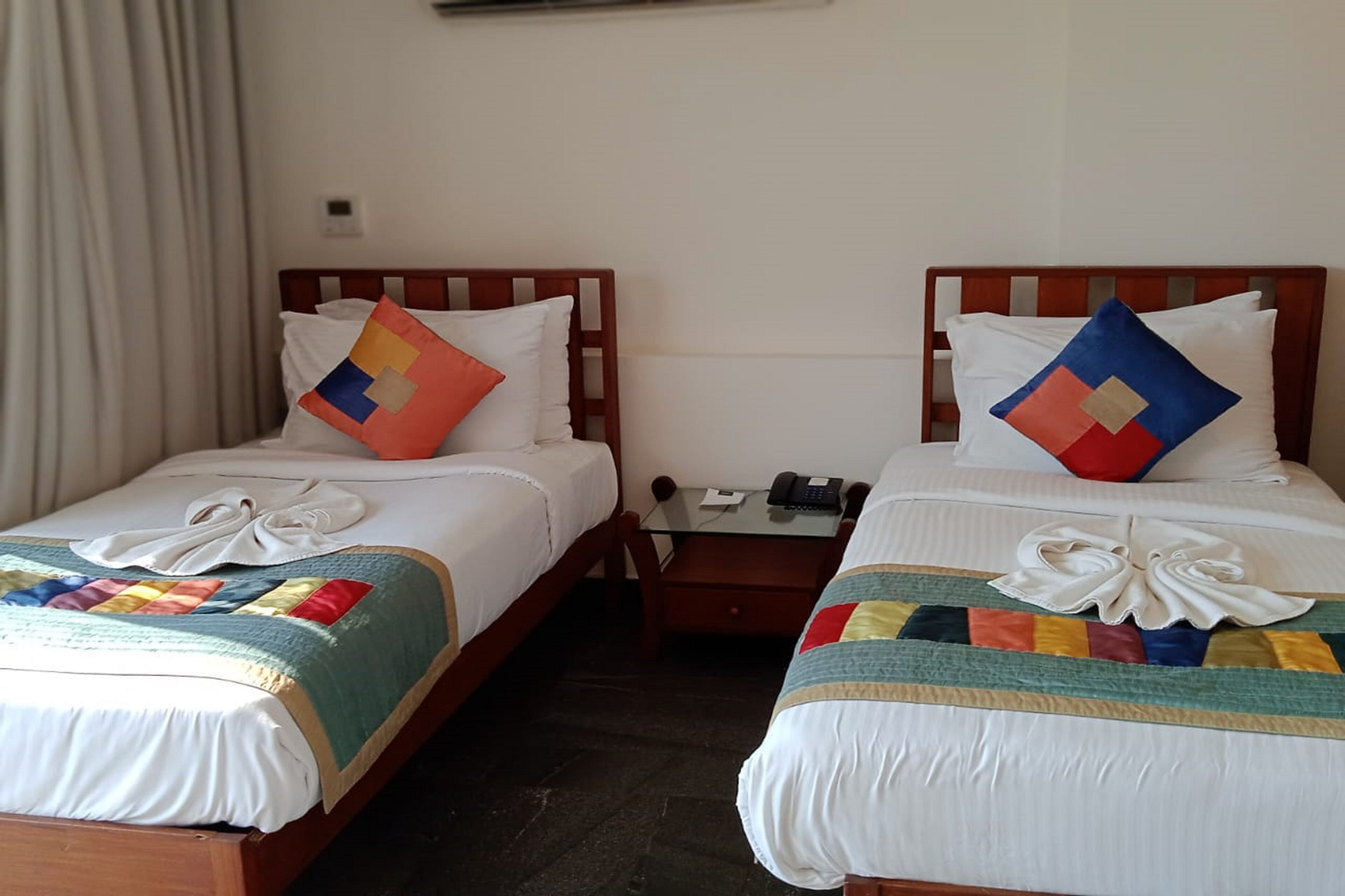 Duas camas de solteiro com lençóis brancos, cobertores coloridos e almofadas decorativas. Cada cama possui uma mesinha de cabeceira com telefone e luminária.