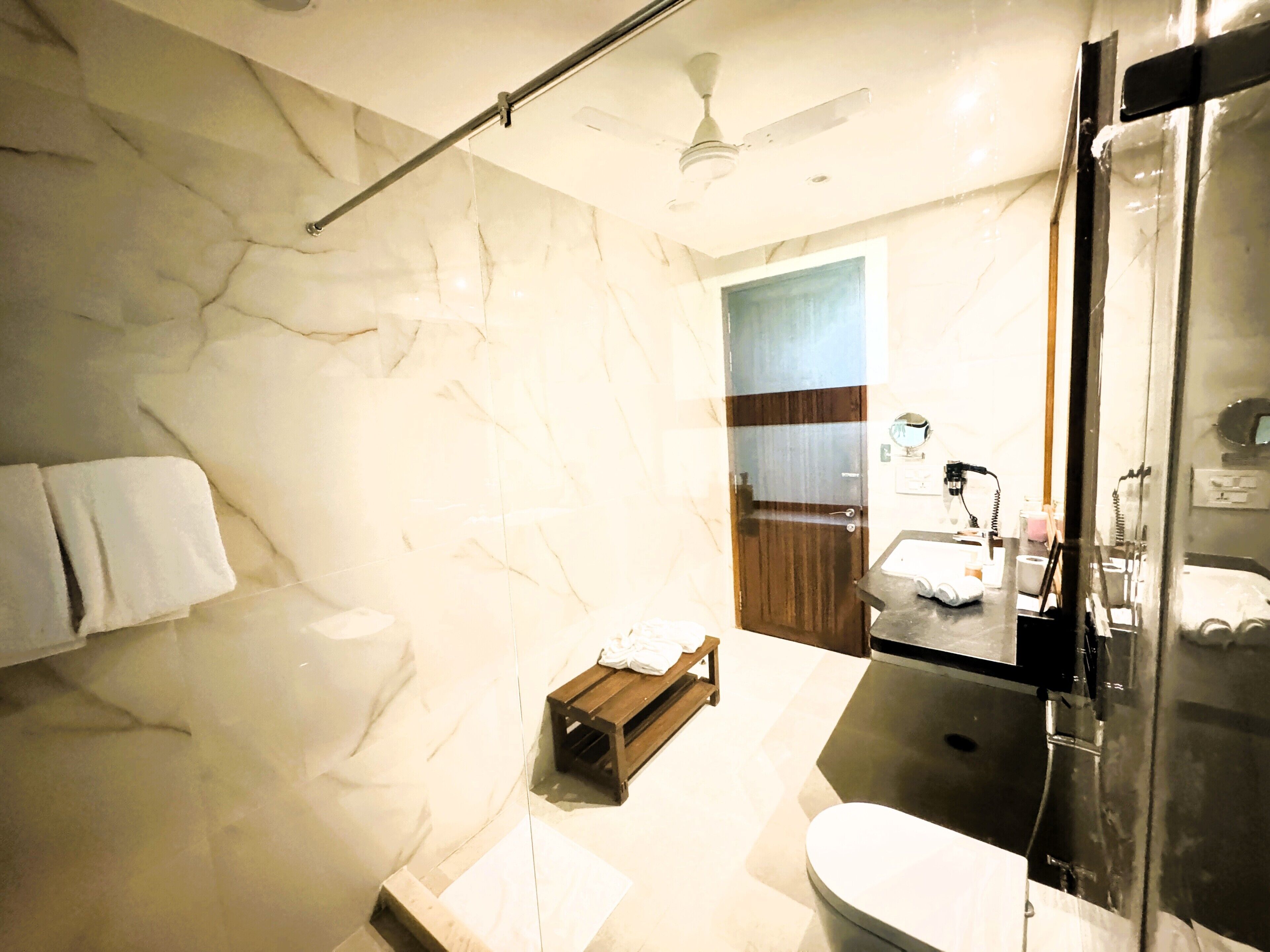 A modern bathroom with a marble wall, a ceiling fan, a wooden door, and a glass shower enclosure.
