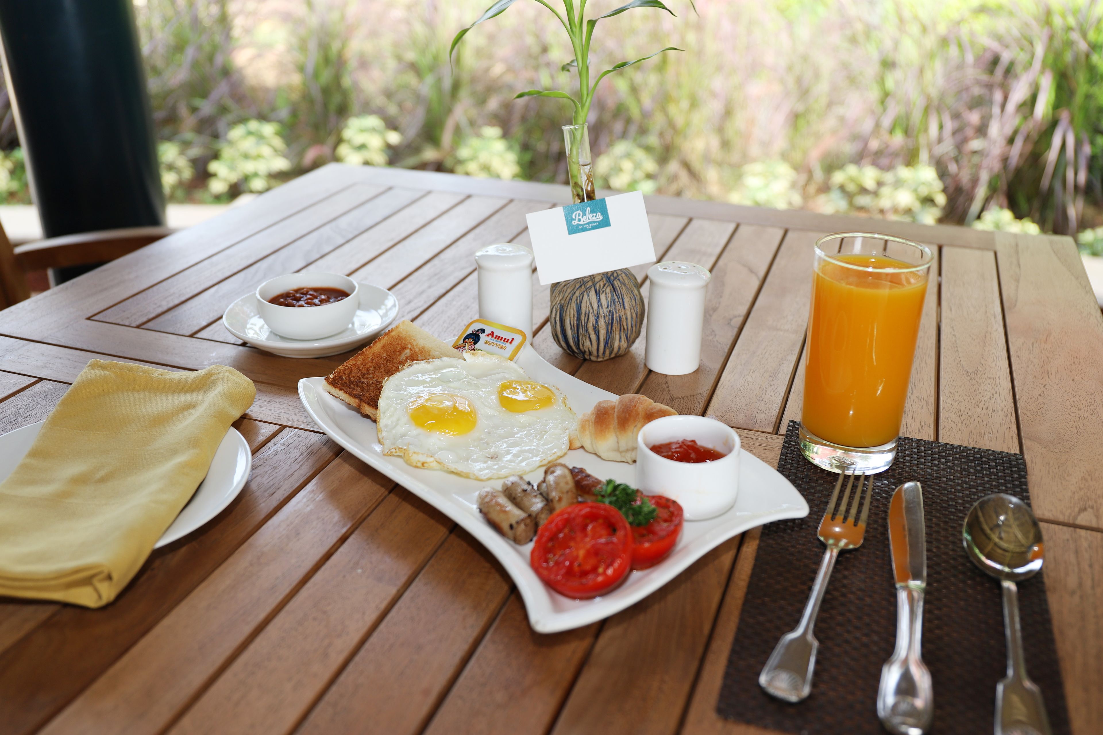 A breakfast table with a plate of eggs, toast, and tomatoes, a glass of orange juice, and a small plant with a name tag.