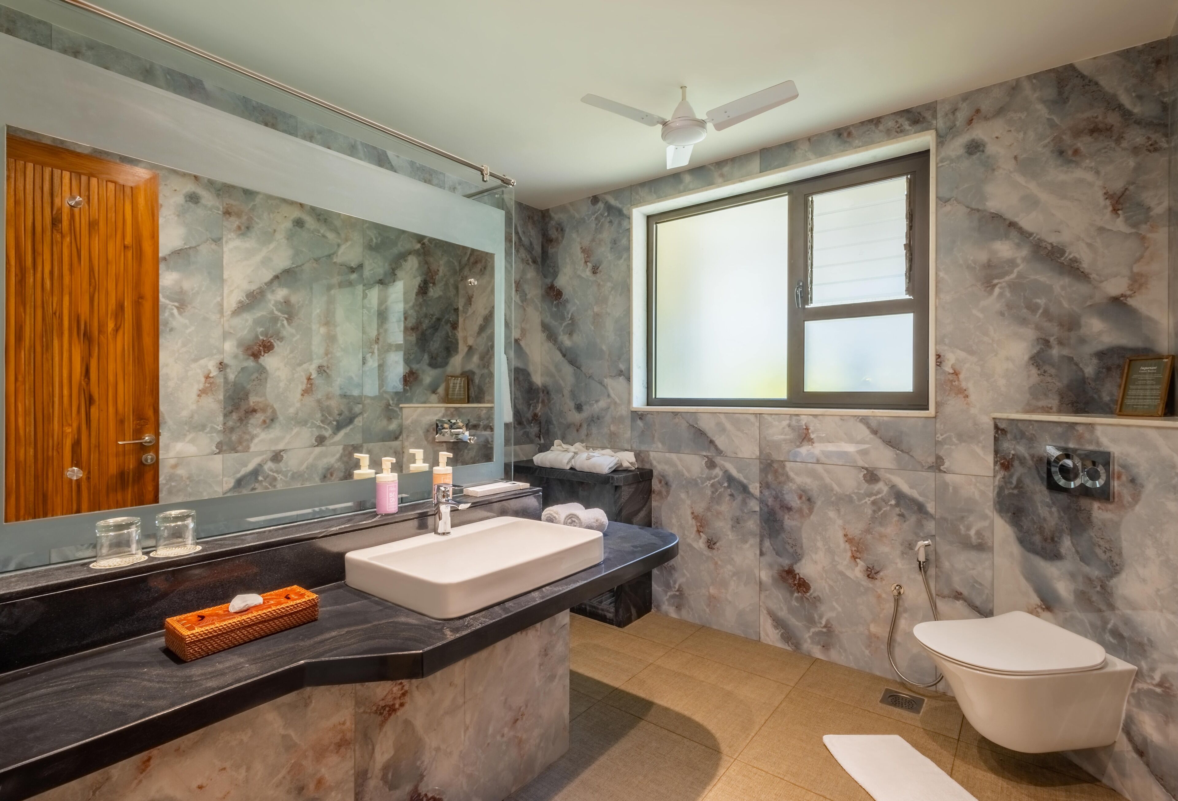A modern bathroom with a marble wall, a large mirror, a freestanding sink, a toilet, and a ceiling fan.