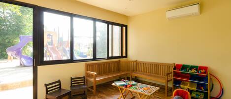 A room with a large window, a wooden bench, a table with toys, and a play area visible outside.