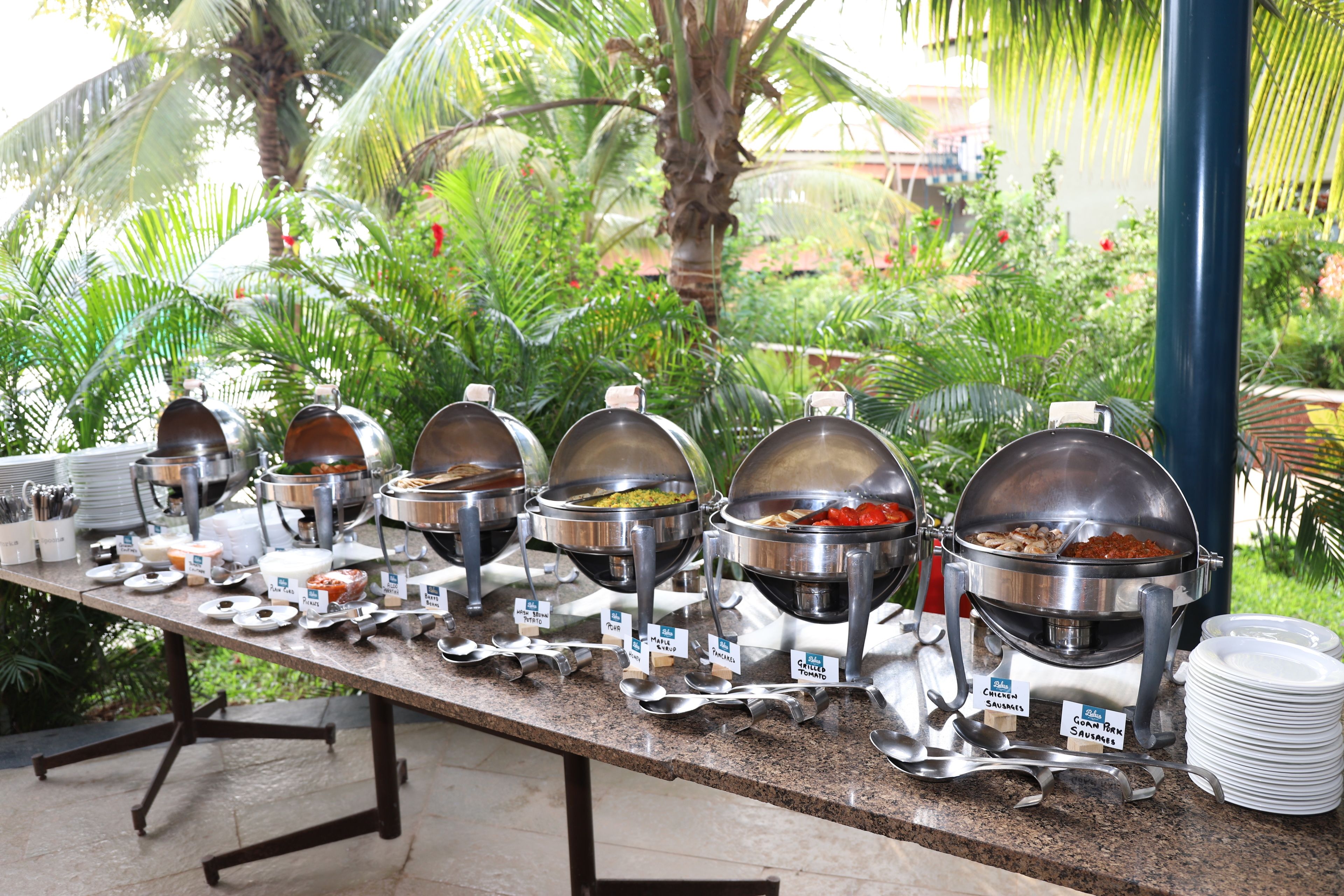 Una mesa de buffet con diversos alimentos y utensilios de servicio, con abundante follaje y una palmera de fondo.