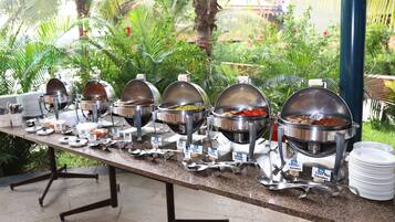 Una mesa de buffet con diversos alimentos y utensilios de servicio, con abundante follaje y una palmera de fondo.