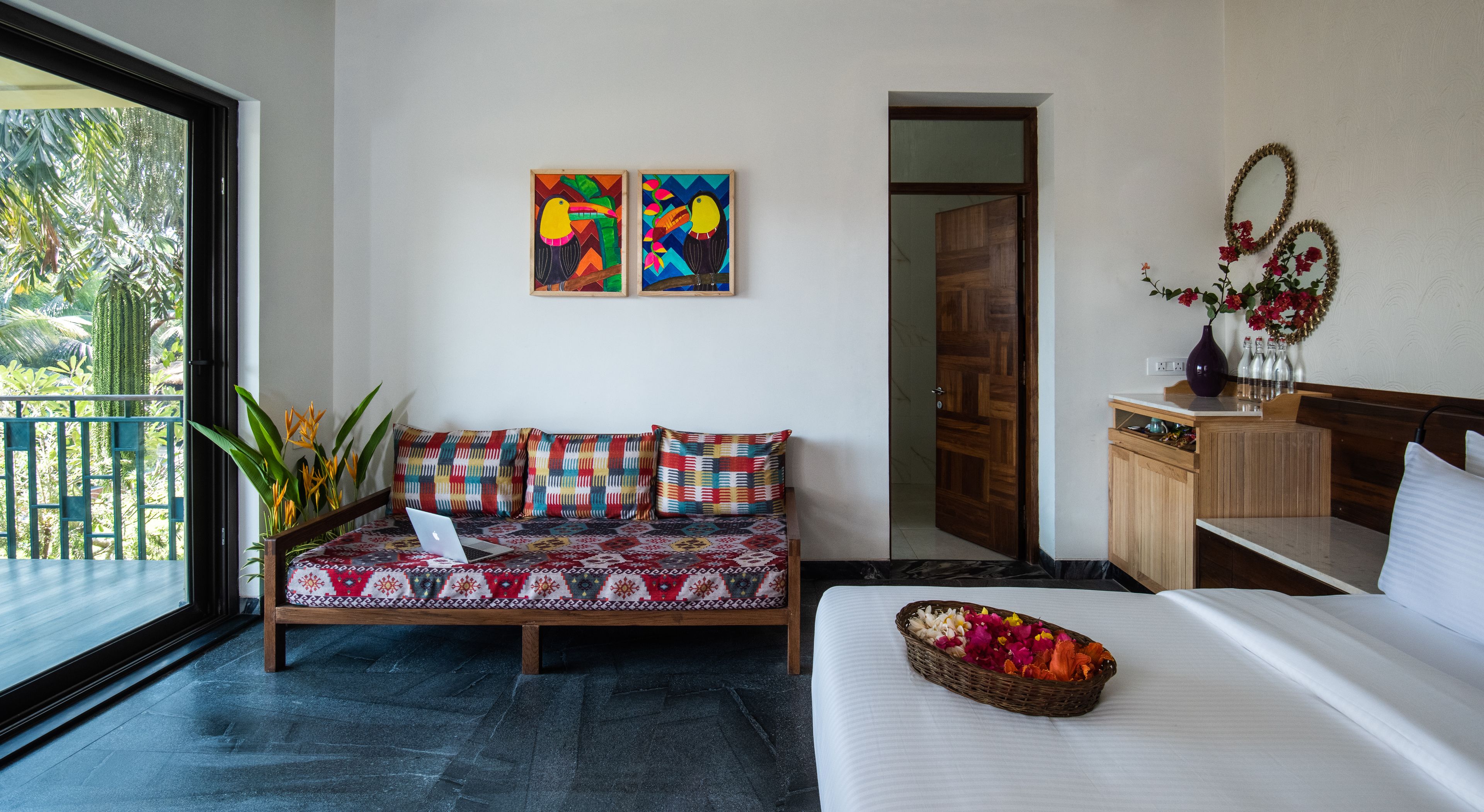 A room with a bed, a sofa, a desk, and a window with a view of greenery. There are two paintings on the wall and a vase with flowers on the desk.