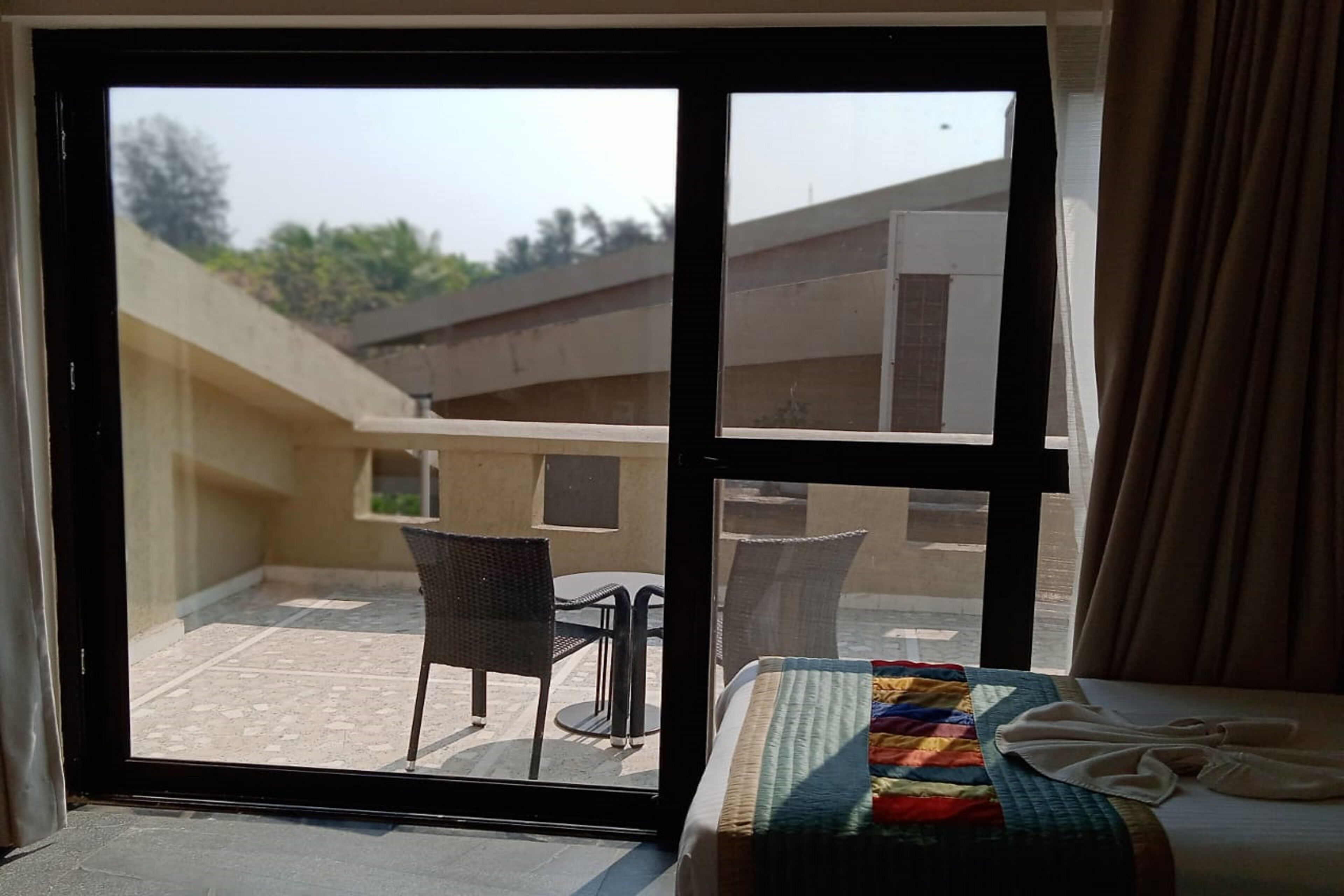 A hotel room with a bed, a window with a view of the outdoor area, and a balcony with chairs and a table.