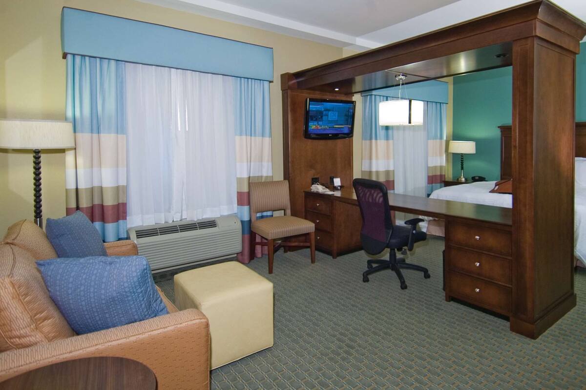 Habitación de hotel con cama, escritorio, silla, sofá, televisor y un espejo grande.