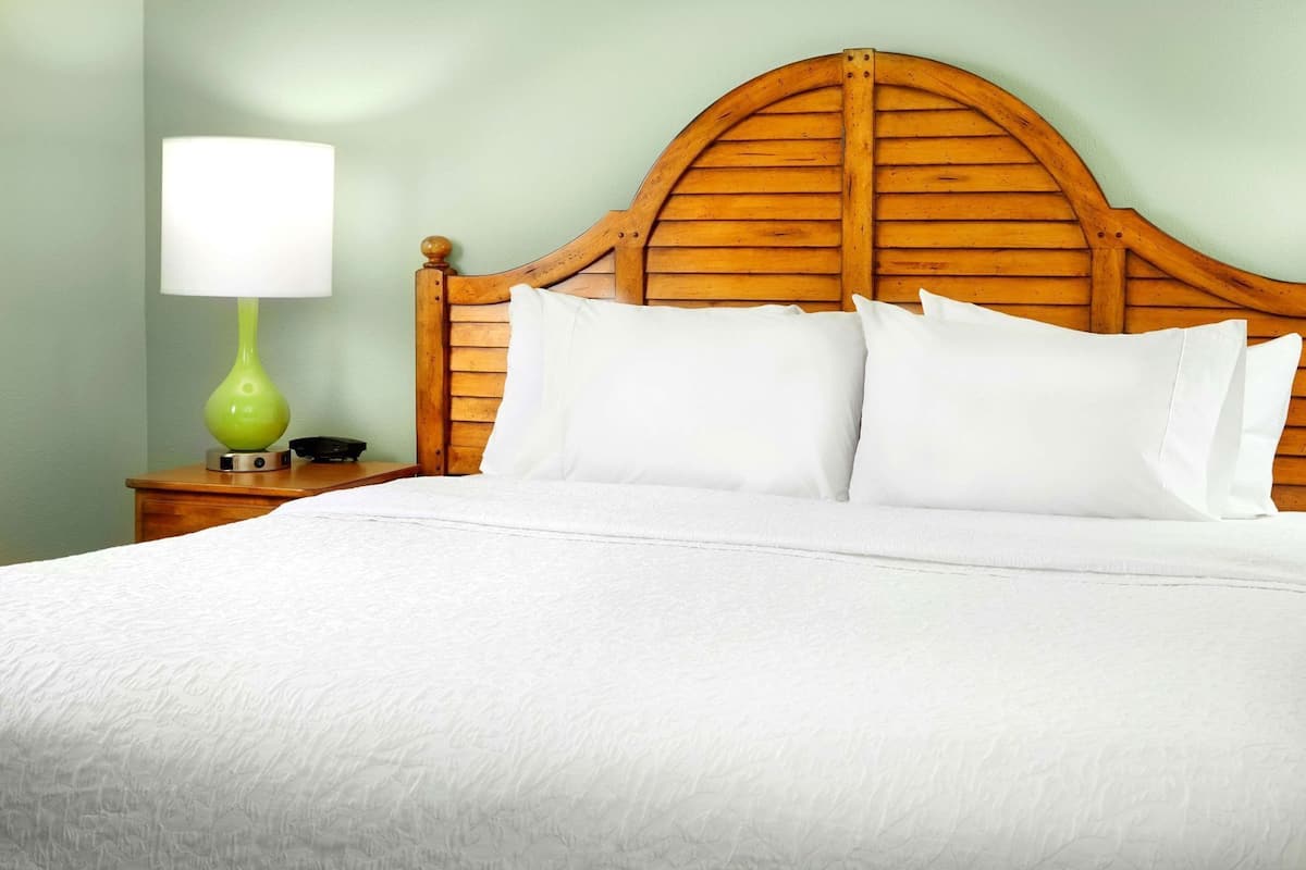 Uma cama com cabeceira de madeira, roupa de cama branca e abajur verde sobre um criado-mudo de madeira.