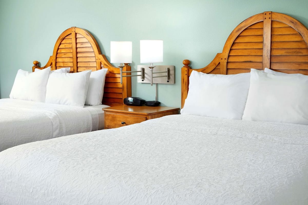 Quarto de hotel com duas camas, cabeceiras de madeira, roupa de cama branca, mesa de cabeceira com abajur e luminária na parede.