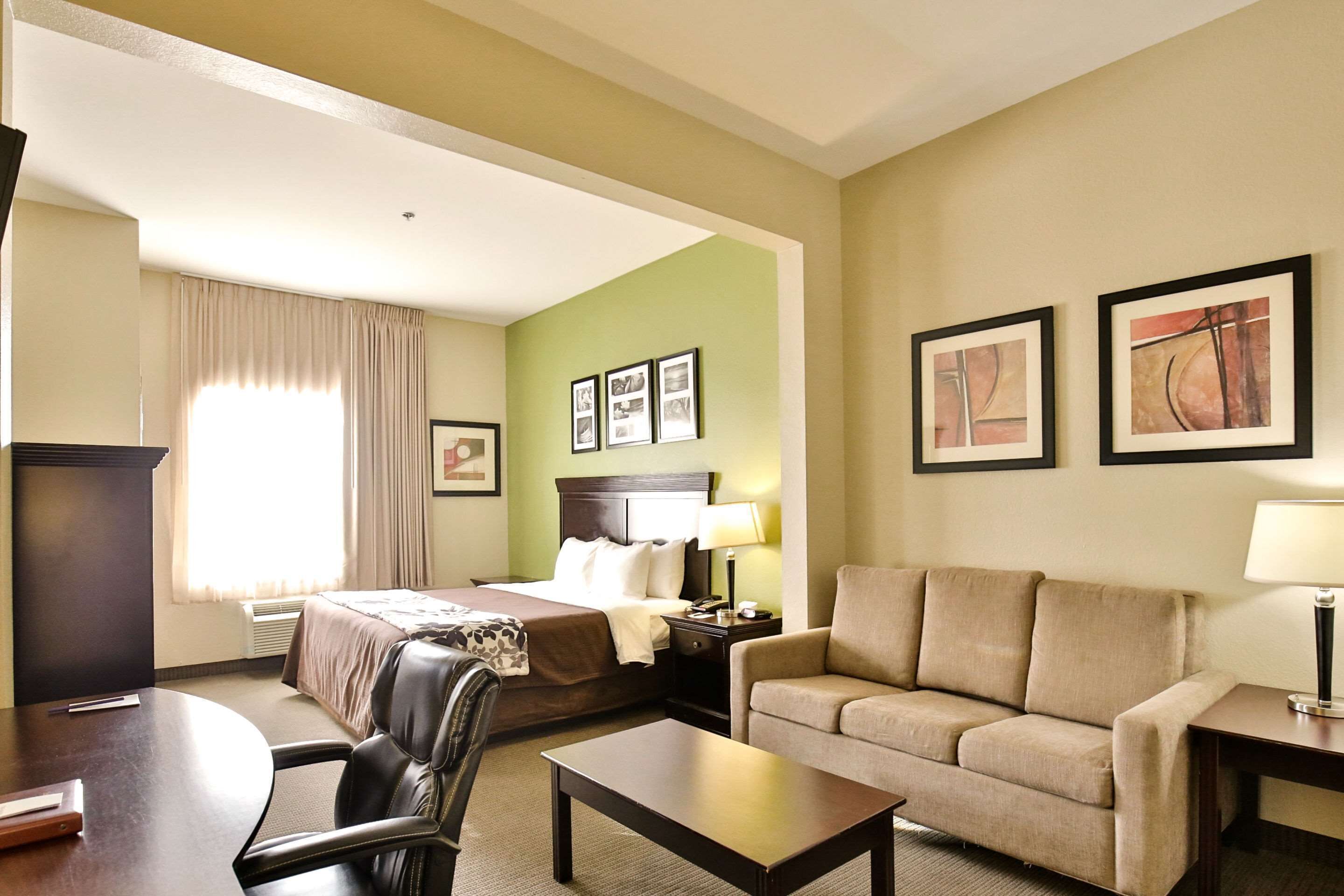 A hotel room with a bed, a sofa, a desk, and a chair. There are framed pictures on the wall and a window with curtains.