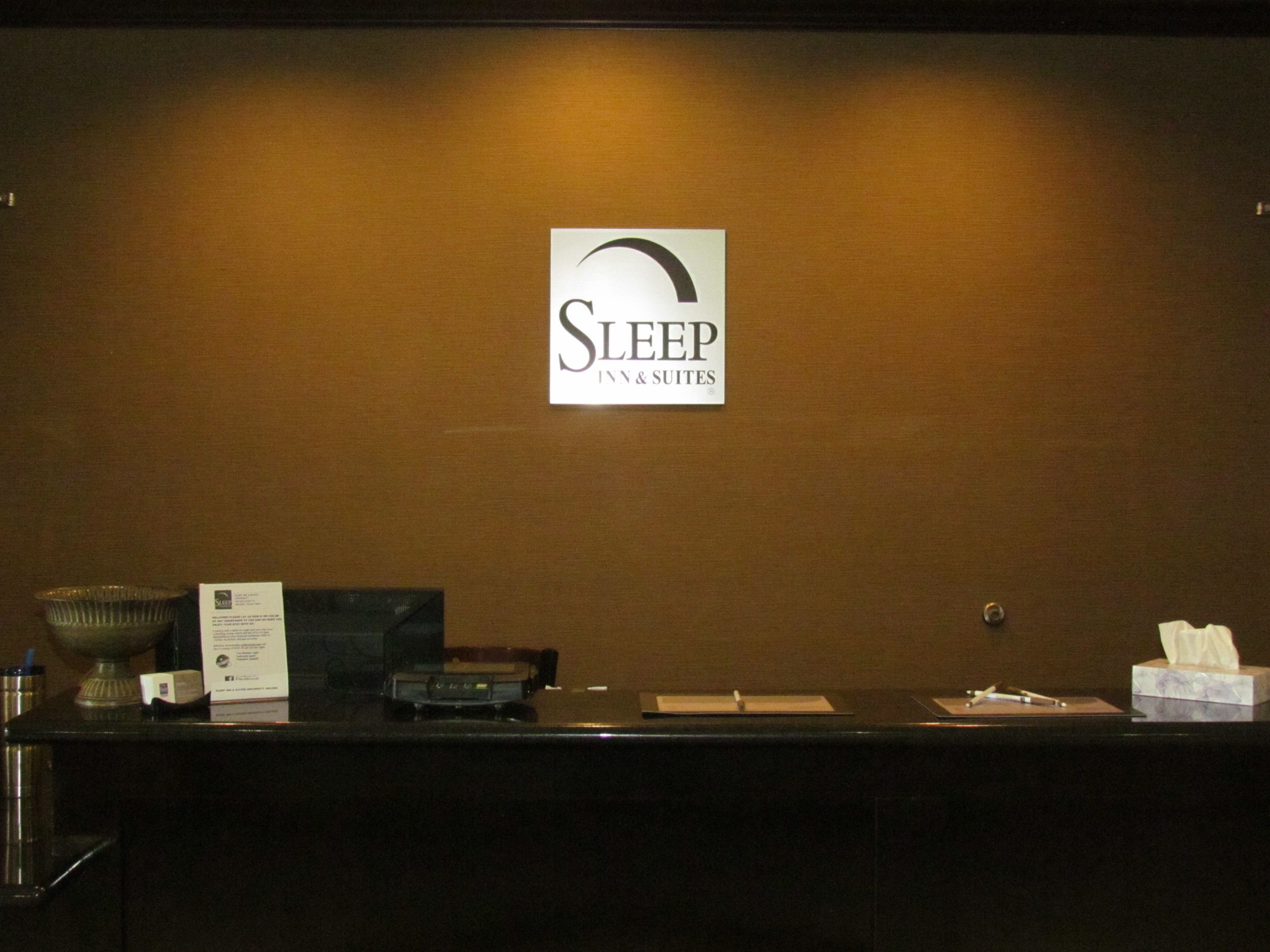 A reception desk with a sign for Sleep Inn & Suites, a glass container, a box, a tissue box, and a pen.