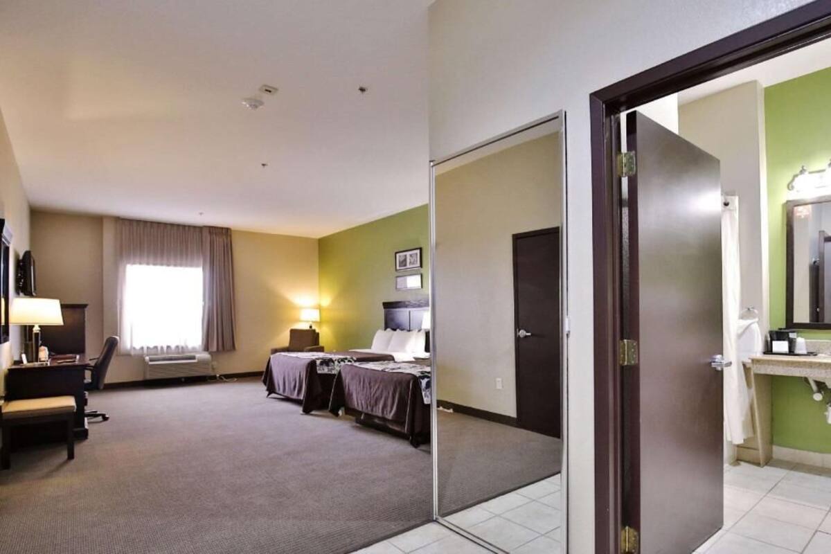Une chambre d’hôtel avec deux lits, un bureau, une chaise, une fenêtre avec des rideaux, une salle de bain avec un miroir et une porte.