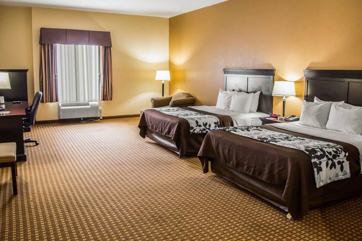 Une chambre d’hôtel avec deux lits, un bureau, une chaise, une lampe et une fenêtre avec des rideaux.