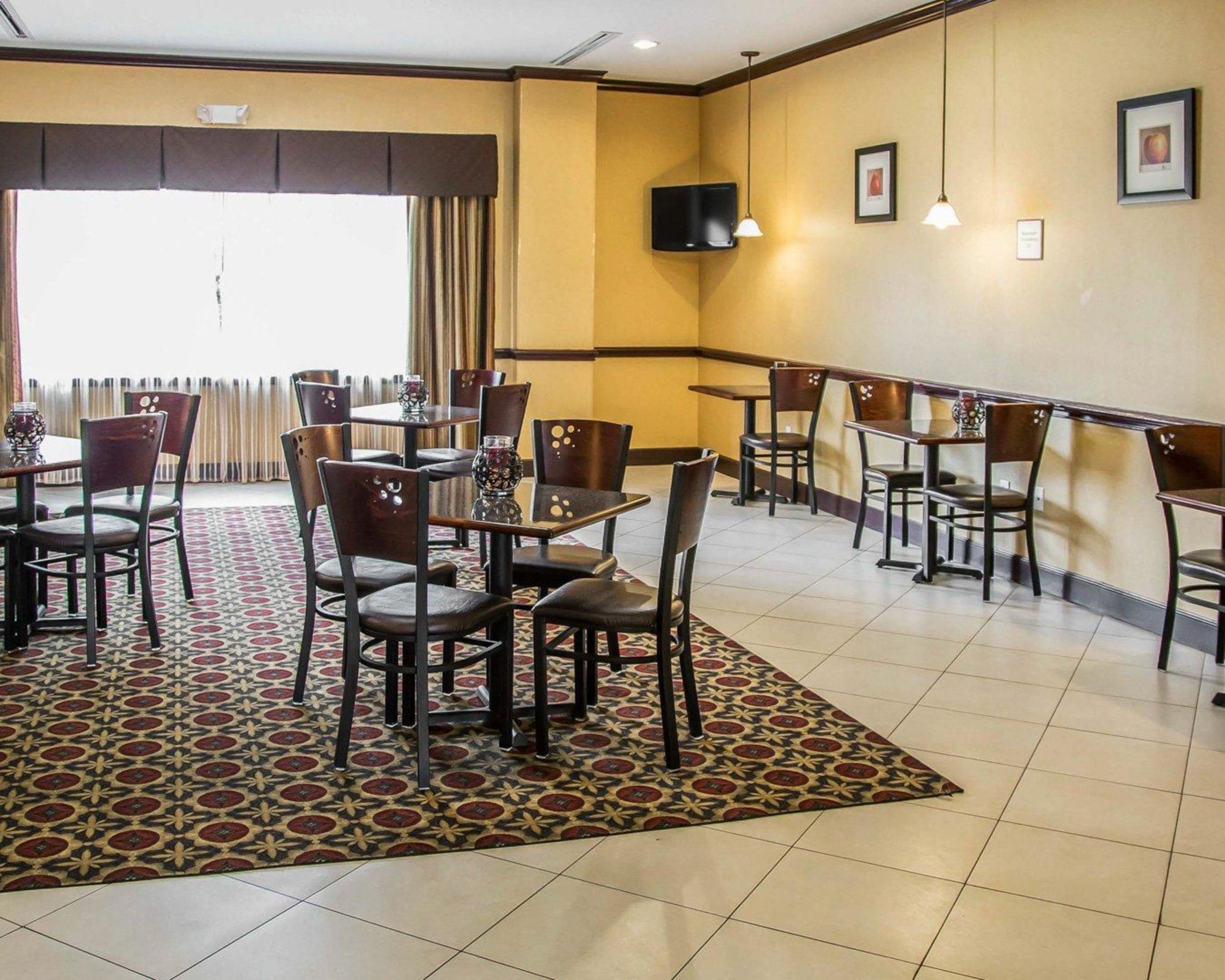 A hotel dining area with multiple tables and chairs, a patterned rug, and a television mounted on the wall.