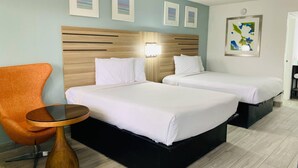 Habitación de hotel con dos camas, cabecera de madera, una mesita, una silla naranja y obras de arte en las paredes.