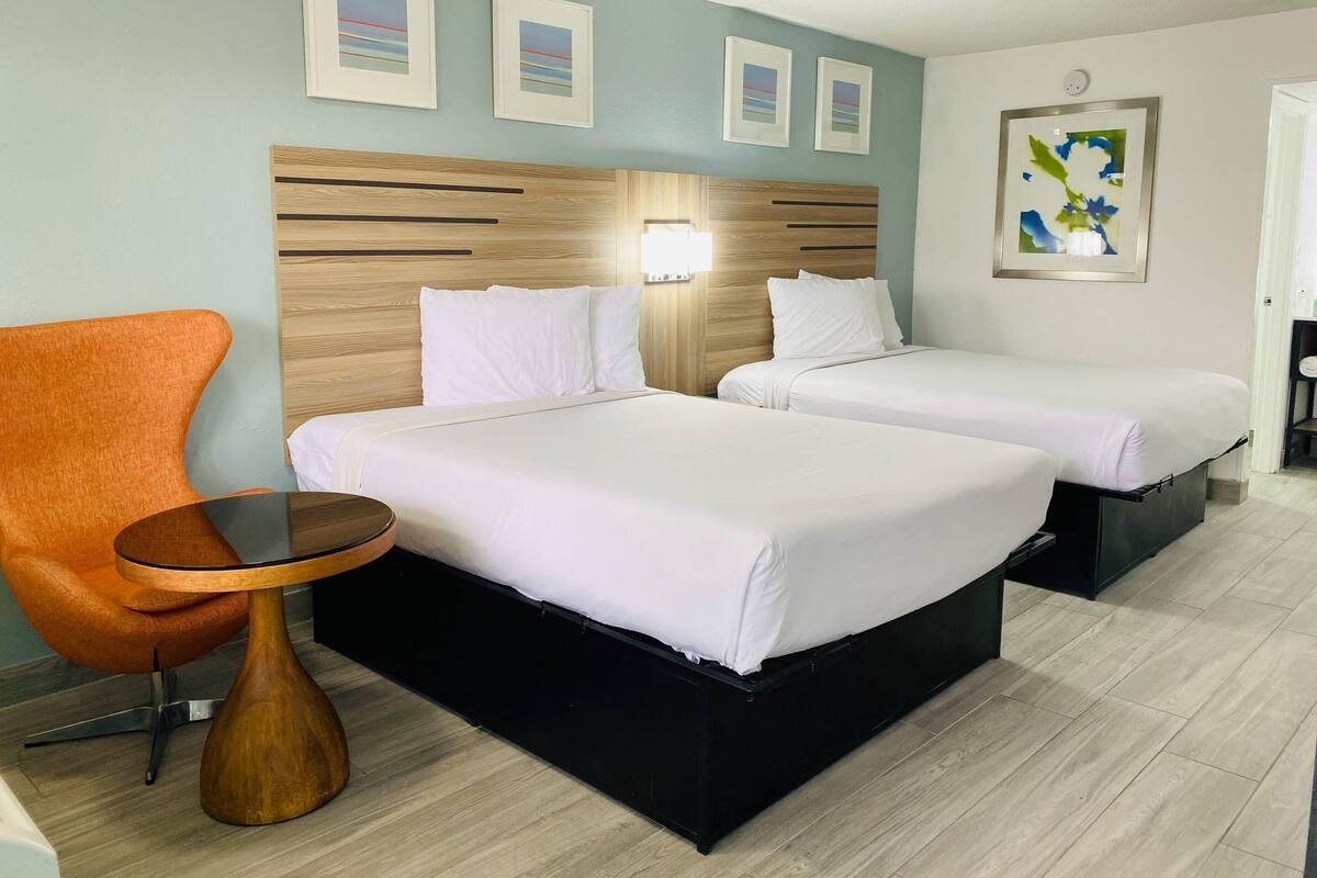 Habitación de hotel con dos camas, cabecera de madera, una mesita, una silla naranja y obras de arte en las paredes.