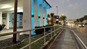 Un edificio moderno con columnas iluminadas, un letrero de estacionamiento por 15 minutos, y una vista de la calle con palmeras y vehículos.