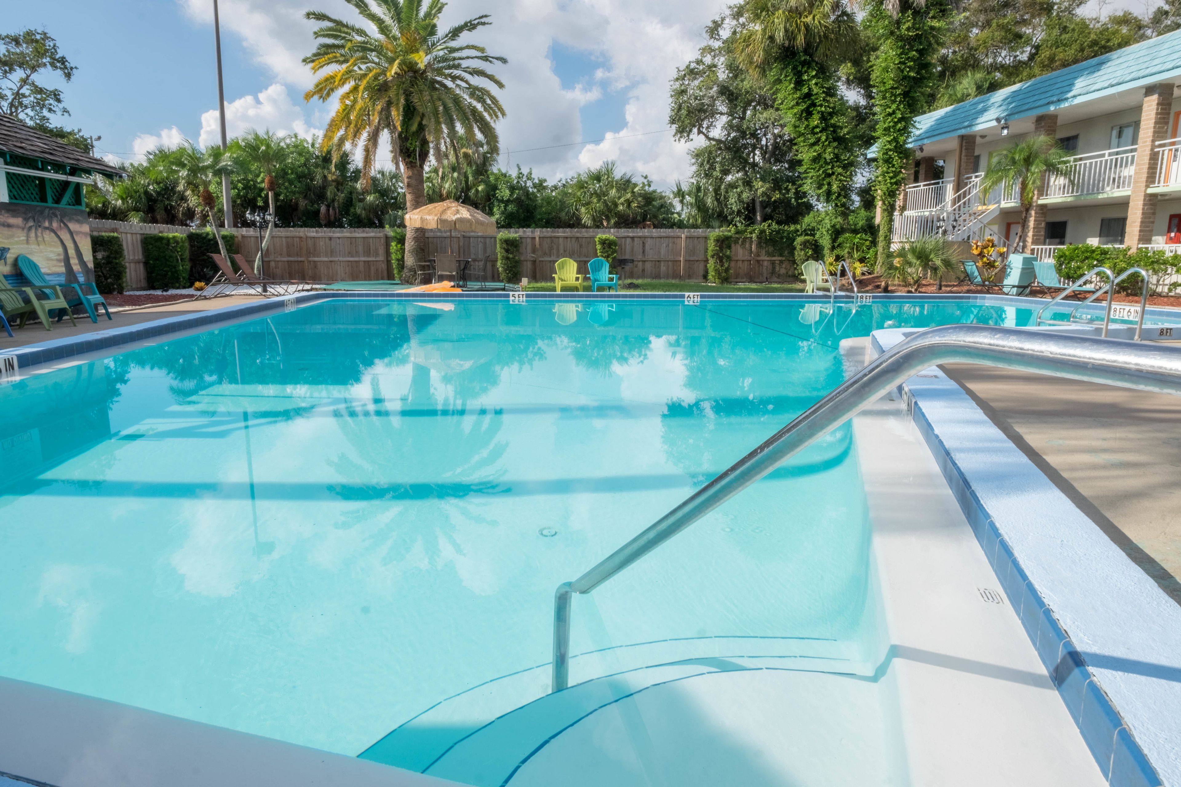 Una piscina azul cristalina con un deck de madera y sillas de descanso, rodeada de palmeras y un edificio con techo azul.