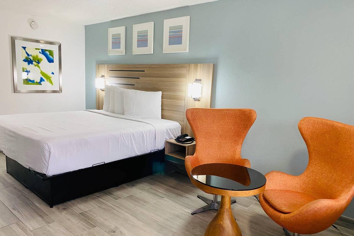 Habitación de hotel con cama, dos sillones naranjas, una mesita redonda y cabecera con lámpara.