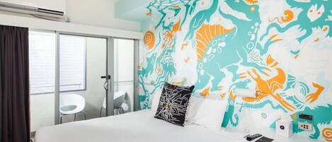 Une chambre d’hôtel avec un lit, une table de chevet blanche, un mur orné d’une fresque colorée, une fenêtre avec des rideaux et une porte.